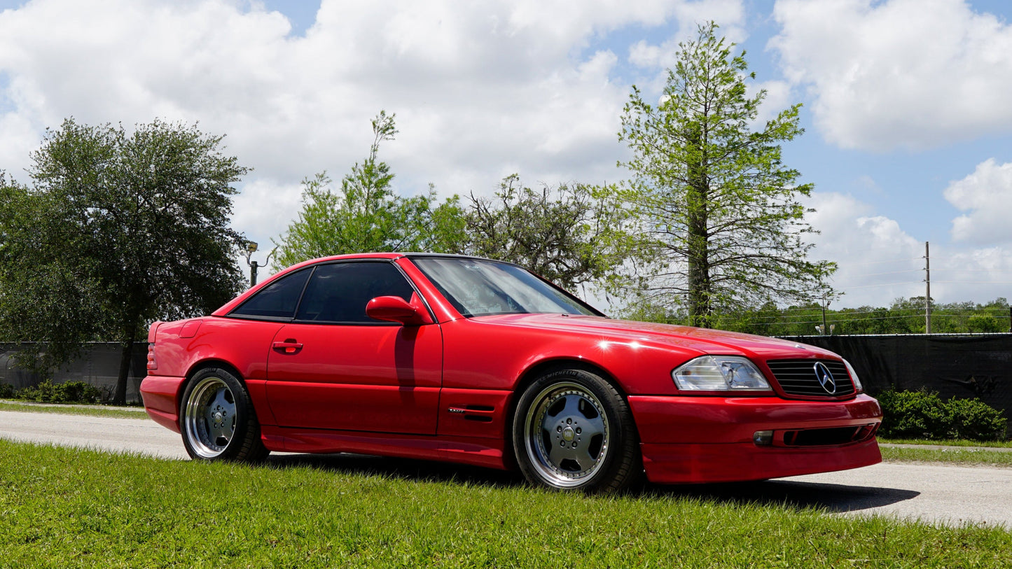 Red R129 Mercedes AMG Sport car parked on a grassy area with trees and a blue sky in the background