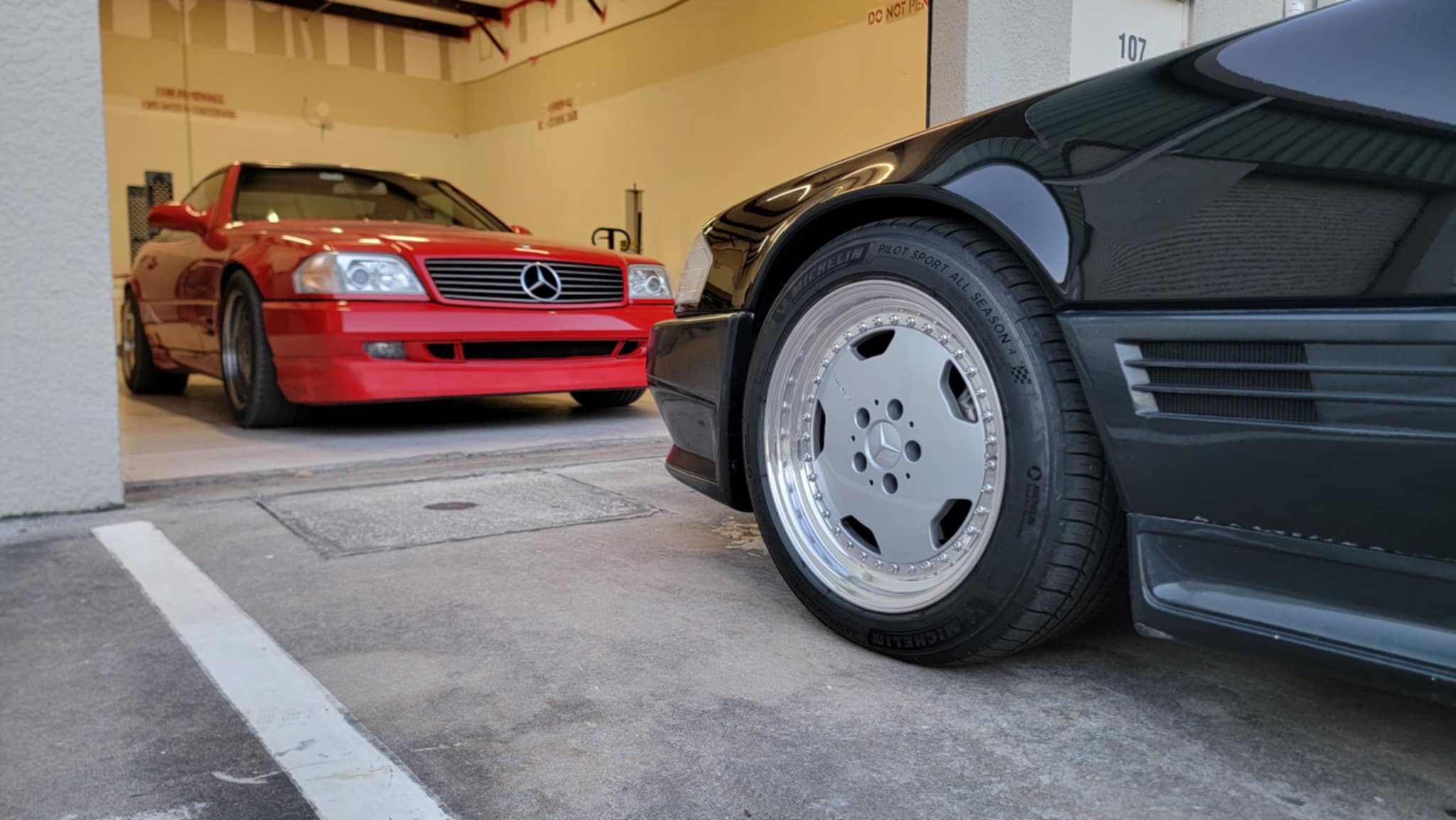 Two cars, a magma red Mercedes R129 and a black AMG 1 R129 Aero sports car, in a garage.